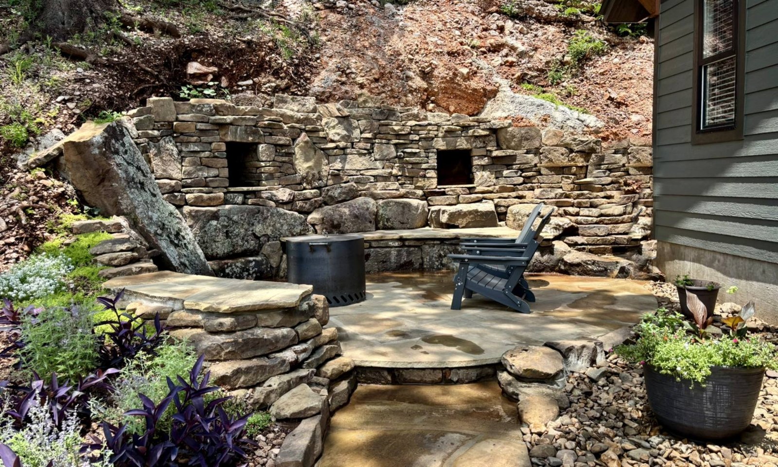 The custom firepit patio: a flagstone circle set into the hillside, with a dry-stacked stone wall, a curved stone bench and a smokeless firepit
