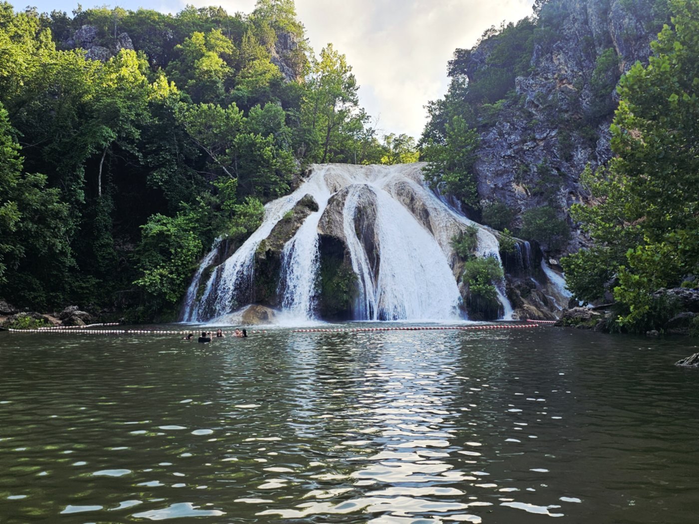 Turner Falls in daylight, water spreading down the rock face into the green swimming pool with people in the water below