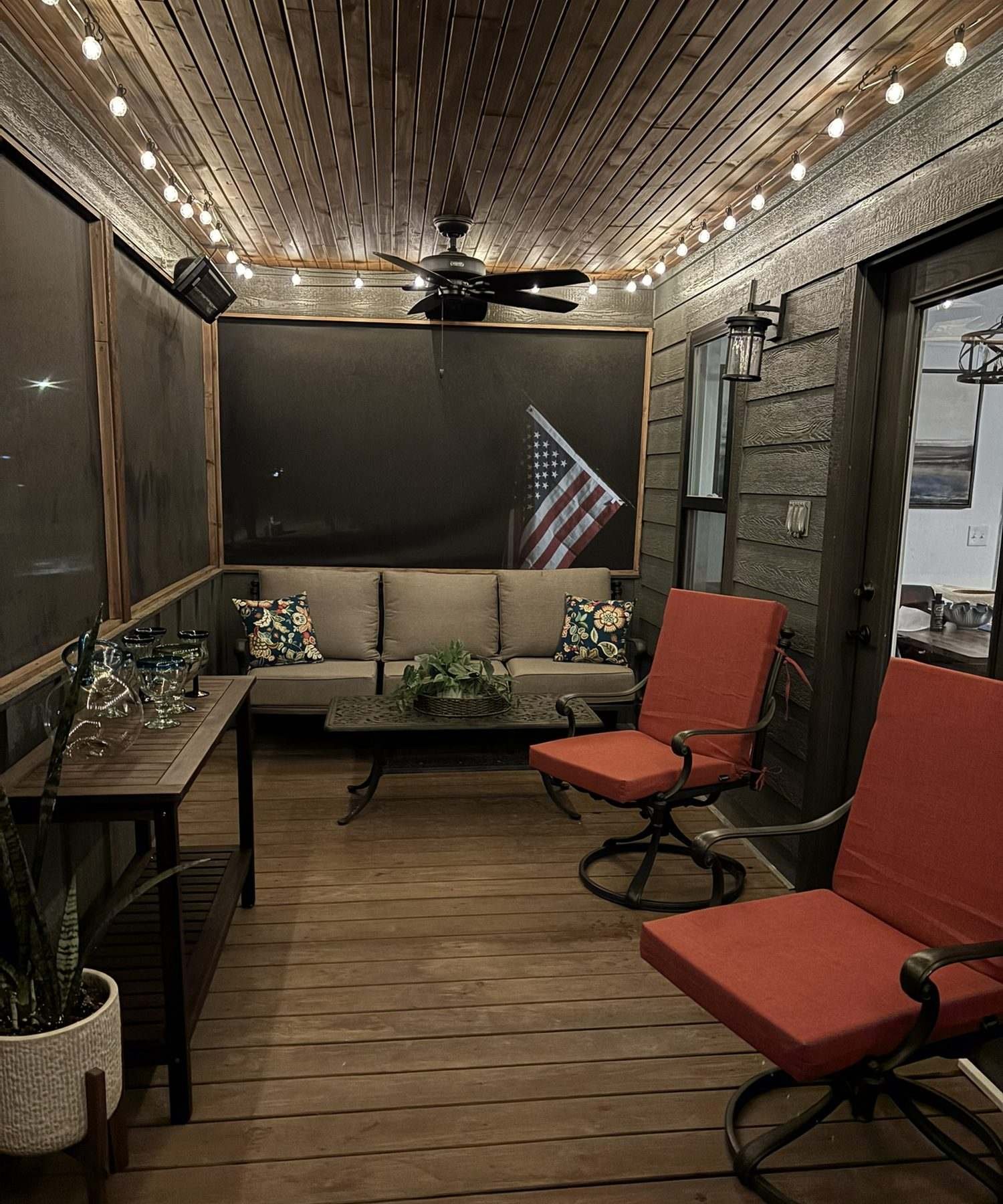 The screened porch at night under warm string lights, with cushioned seating and a ceiling fan